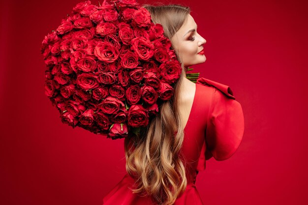 Over shoulder studio portrait of gorgeous young brunette with bright lips in red dress holding big bouquet of red roses and smiling at camera over red background Isolate on red
