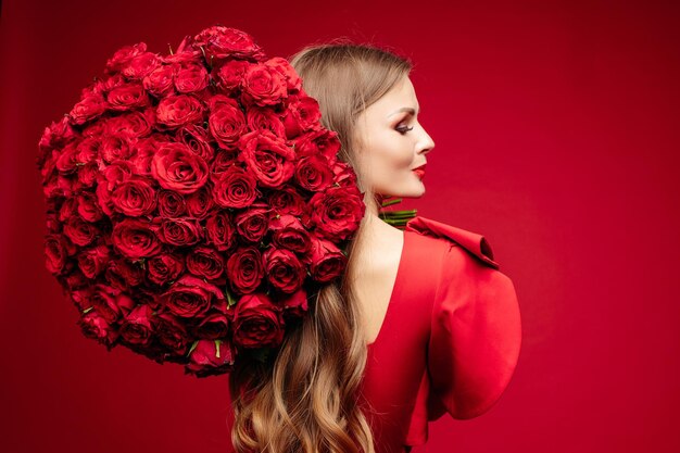 Over shoulder studio portrait of gorgeous young brunette with bright lips in red dress holding big bouquet of red roses and smiling at camera over red background Isolate on red