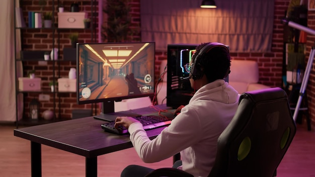 Over shoulder view of african american gamer using pc gaming setup relaxing playing first person shooter in multiplayer. Man streaming while explaining gameplay in online action game talking to team.