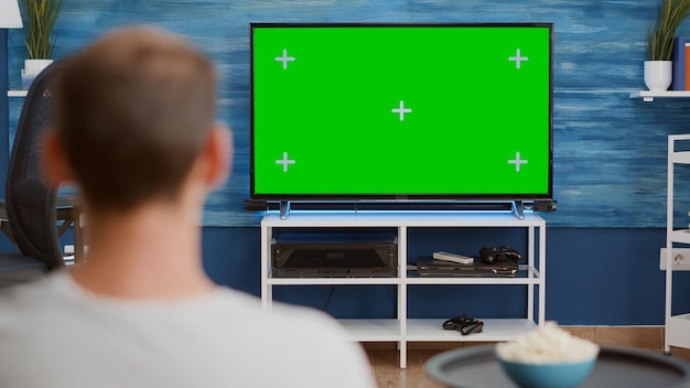 Over shoulder view of man watching movie on tv with green screen relaxing with bowl of popcorn sitting on couch. Back view of person relaxing on sofa in front of television mockup with chroma key disp