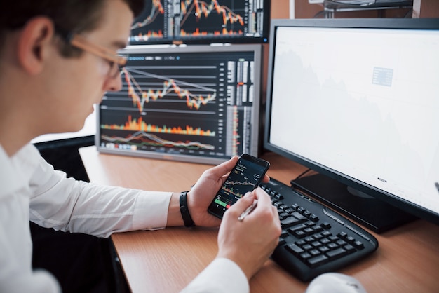 Over the shoulder view of and stock broker trading online while accepting orders by phone. Multiple computer screens ful of charts and data analyses in background