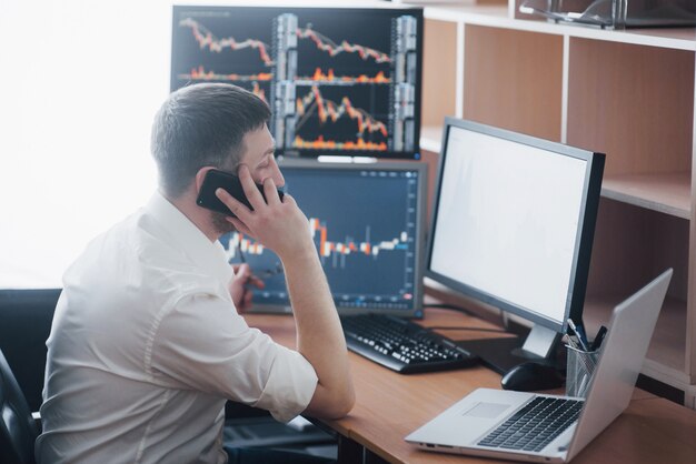 over the shoulder view of and stock broker trading online while accepting orders by phone. multiple computer screens ful of charts and data analyses in background