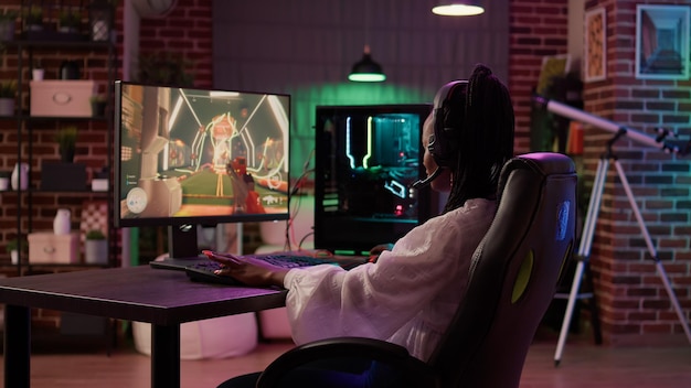 Over shoulder view of woman streaming first person shooter doing victory hand gesture after win in tournament. African american gamer girl surprised after winning online competition on gaming pc.