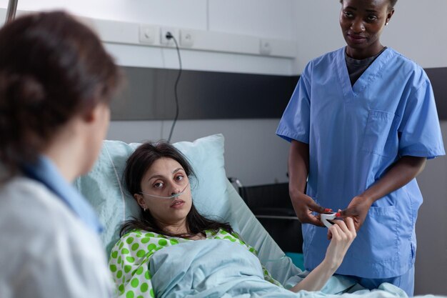 Over shoulder view of woman with low spo2 saturation recieving oxygen recovering in hospital bed looking at doctor providing medical care. Nurse attending patient with nasal cannula in hospital ward.