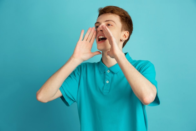 Shouting, calling. Caucasian young man's modern portrait isolated on blue  wall, monochrome. Beautiful male model. Concept of human emotions, facial expression, sales, ad, trendy.