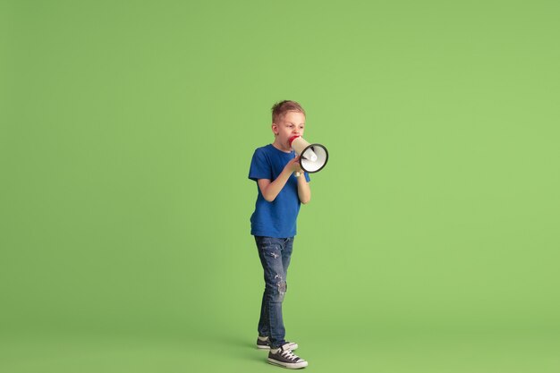 叫び、呼びかけ。緑の壁で遊んで楽しんでいる幸せな少年。明るい布の白人の子供は、遊び心のある、笑顔に見えます。教育、子供時代、感情、表情の概念。