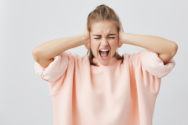 Shouting emotional in despair young caucasian woman, with open mouth and closed eyes, screaming, closing her ears as refusing to listen bad news. Face expression, reaction and negative emotions.