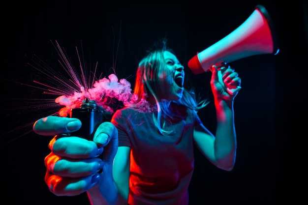 Shouting with megaphone. Young woman with smoke and neon light on black background. Highly tensioned, wide angle, fish eye view. 