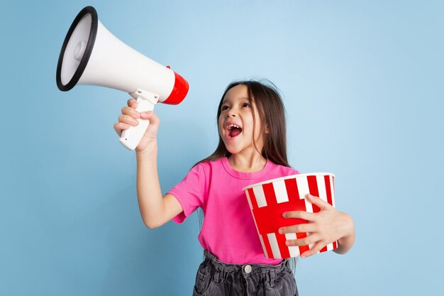 Shouting with popcorn. Caucasian little girl's portrait on blue wall. Beautiful female model in pink shirt.