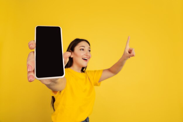 Showing blank phone's screen. Caucasian woman's portrait isolated on yellow studio background. Beautiful brunette model in casual. Concept of human emotions, facial expression, sales, ad, copyspace.