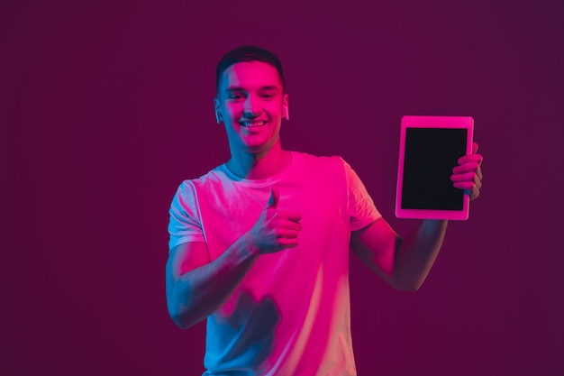 Showing blank screen. Caucasian man's portrait isolated on pink-purple  wall in neon light. Male model with devices. Concept of human emotions, facial expression, Copyspace.
