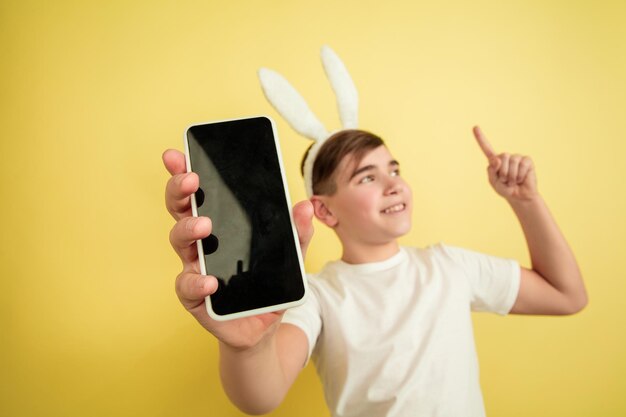 Showing blank screen, pointing up. Caucasian boy as an Easter bunny on yellow background. Happy easter greetings.