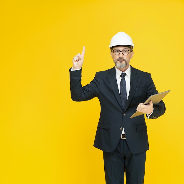 Showing finger up middle aged business man in helmet or hard hat with tablet tc in hand looking at camera isolated on yellow background Handsome mature businessman in black suit Business concept