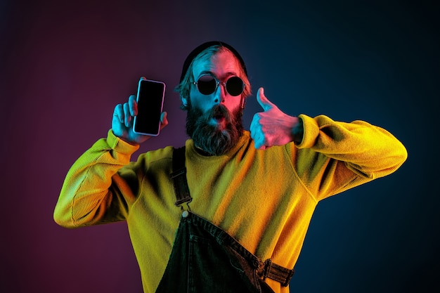 Showing phone's blank screen. Caucasian man's portrait on gradient studio background in neon light. Beautiful male model with hipster style. Concept of human emotions, facial expression, sales, ad.