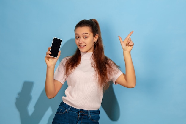Показываю экран телефона, указывая вверх. Портрет кавказской девочки-подростка на синем фоне. Красивая модель в повседневной одежде. Понятие человеческих эмоций, выражения лица, продаж, рекламы. Copyspace. Выглядит счастливым.