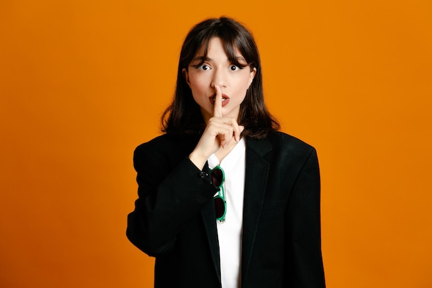 Showing silence gesture young beautiful female wearing black jacket isolated on orange background