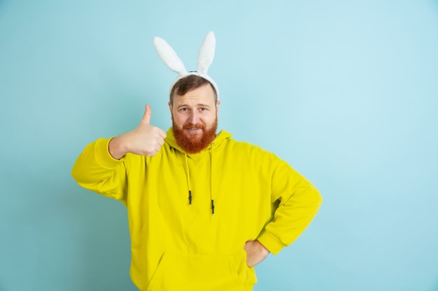 Showing thumb up. Caucasian man as an Easter bunny with bright casual clothes on blue studio background. Happy easter greetings. Concept of human emotions, facial expression, holidays. Copyspace.