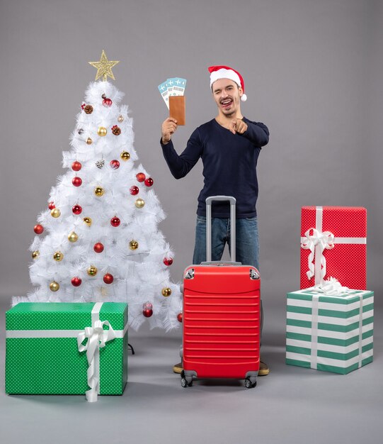 흰색 크리스마스 트리 주위에 그의 여행 티켓을 보여주는 빨간 가방으로 혀 남자를 보여주는 회색에 선물