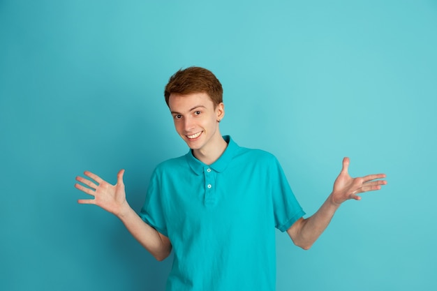 Free photo showing, uncertain. caucasian young man's modern portrait isolated on blue  wall, monochrome. beautiful male model. concept of human emotions, facial expression, sales, ad, trendy.