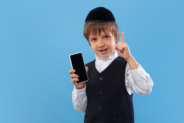 Shows blank phone screen. Portrait of orthodox jewish boy isolated on blue wall. Purim, business, festival, holiday, childhood, celebration Pesach or Passover, judaism, religion concept.
