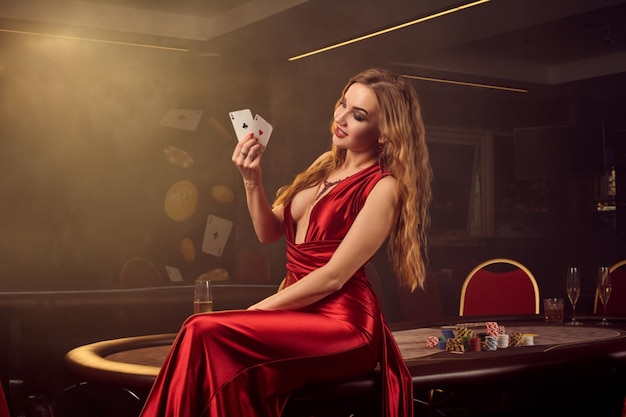Showy blond maiden in a long red satin dress, with two aces in her hand is posing sitting sideways on a poker table in luxury casino. Passion, cards, chips, alcohol, win, gambling - it is a female ent