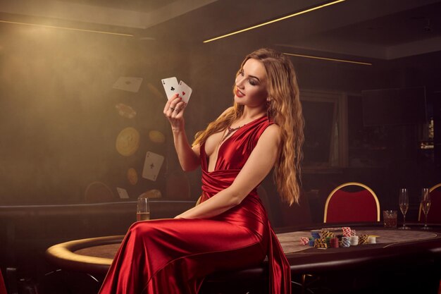 Showy blond maiden in a long red satin dress, with two aces in her hand is posing sitting sideways on a poker table in luxury casino. Passion, cards, chips, alcohol, win, gambling - it is a female ent