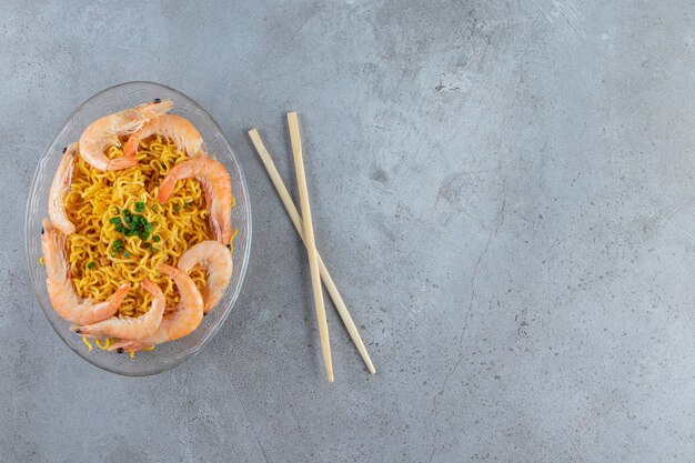 Креветки и лапша на стеклянном блюде рядом с палочками для еды, на мраморном фоне.