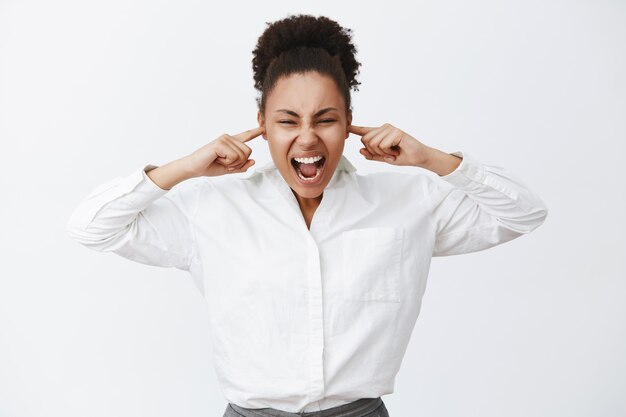 Shut up everybody. Portrait of intense distressed female african-american businesswoman in white shirt, shouting while closing ears with fingers, being pissed and angry, standing in loud place