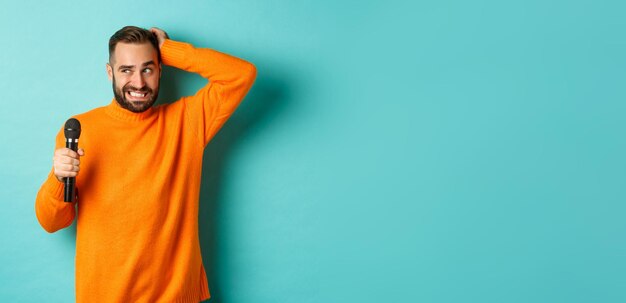 Shy and awkward adult man scratching head holding microphone before singing standing over blue backg