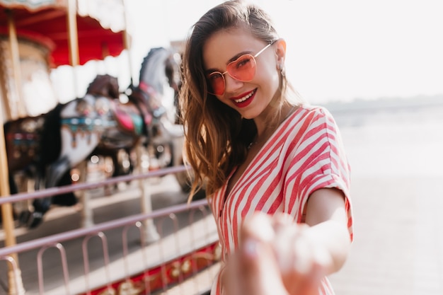 Free photo shy beautiful girl in heart sunglasses standing near carousel. outdoor portrait of blithesome woman in striped attire smiling