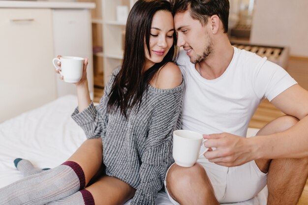 Shy brunette woman in gray sweater and half-hose posing on bed with husband