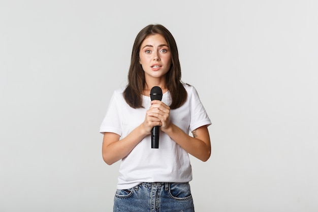 Shy cute brunette girl scared of singing in public, standing with microphone and looking nervous.