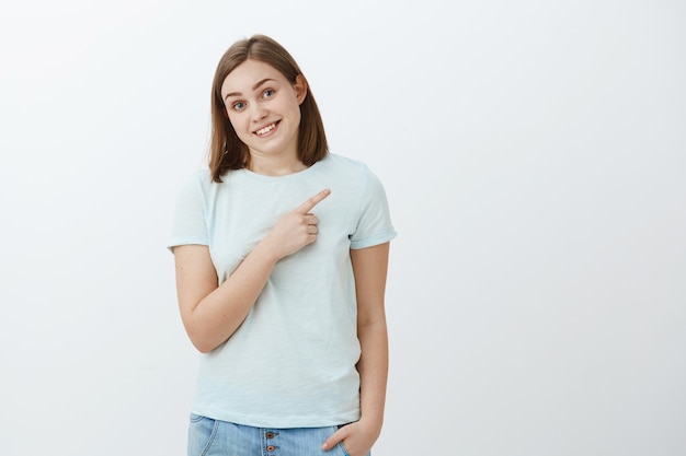 shy cute carefree brunette sportswoman in trendy t-shirt stooping tilting head and smiling friendly and proud pointing at upper right corner showing her medals posing over white wall