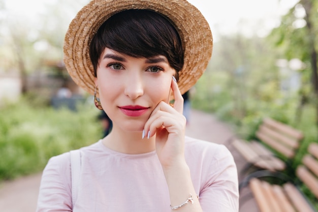 Shy dark-haired lady with red lips wearing silver bracelet touching her face with finger