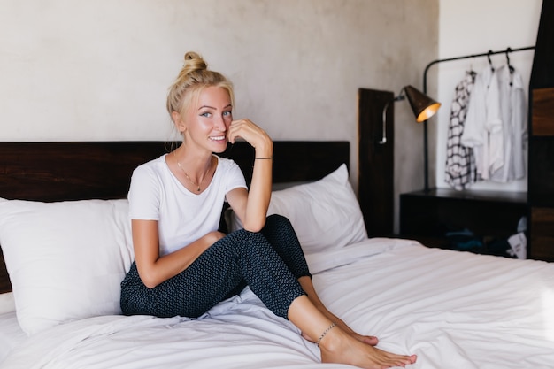 Timida donna bionda con la pelle abbronzata seduta sul letto. foto dell'interno della giovane signora entusiasta che trascorre la mattina in camera da letto.