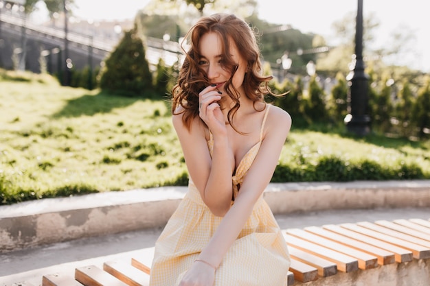 ベンチに座って笑っている巻き毛の髪型の恥ずかしがり屋の生姜の女の子。エレガントな黄色のドレスを着たきれいな女性モデルの屋外写真。