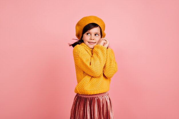 ピンクの壁にポーズをとる恥ずかしがり屋の少女。黄色い服装のかわいい子。