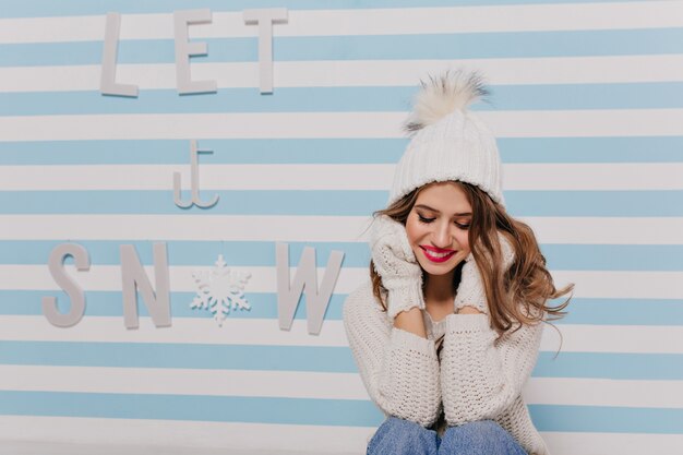 Shy pretty lady in knitted hat and warm sweater sitting modestly with sweet smile