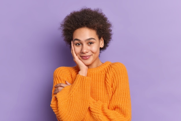 Shy pretty teenage girl with curly hair keeps hand on cheek smiles gently being in good mood wears casual knitted jumper.