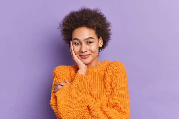 Shy pretty teenage girl with curly hair keeps hand on cheek smiles gently being in good mood wears casual knitted jumper.