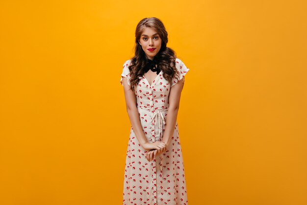 Застенчивая женщина в платье с цветочным принтом позирует на оранжевом фоне. Довольно молодая девушка с фигурной девушкой в стильной одежде, глядя в камеру.