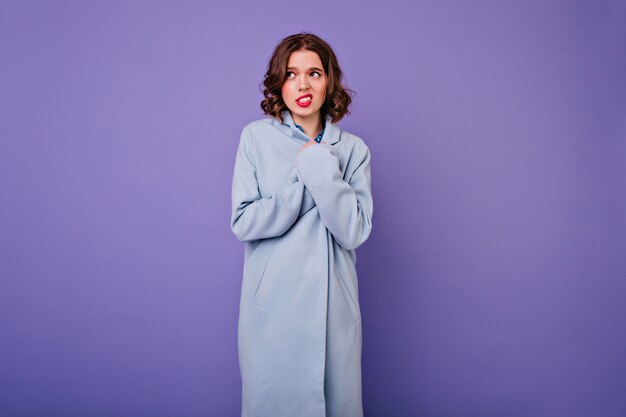 Застенчивая женщина с короткими волнистыми волосами позирует в элегантном длинном пальто. Красивая европейская кудрявая девушка в синем наряде, изолированном на фиолетовой стене.
