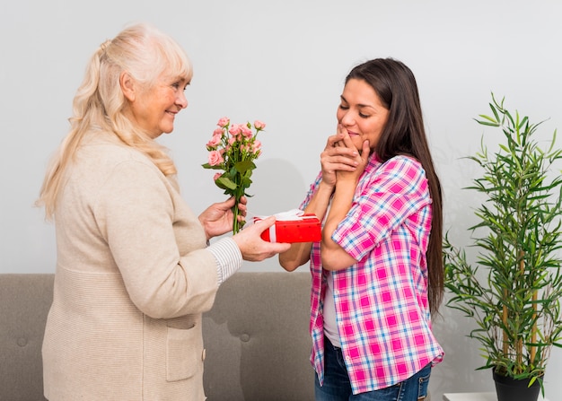 Shy young woman looking at smiling mother holding roses bouquet and gift box