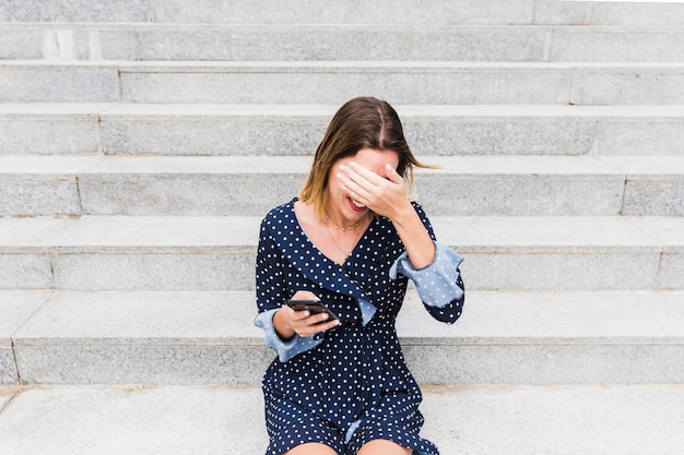 Застенчивая молодая женщина, сидя на лестнице, проведение смартфон