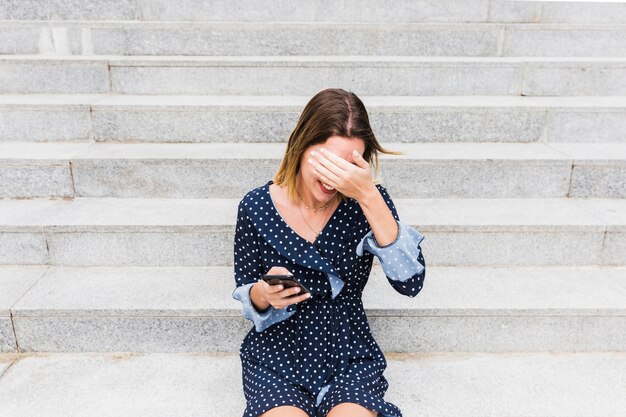 Shy young woman sitting on staircase holding smartphone