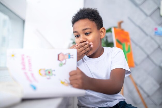 Shyness. Cute dark-skinned school-age boy in tshirt surprised looking at book shyly covering his mouth with hand sitting near window in bright room