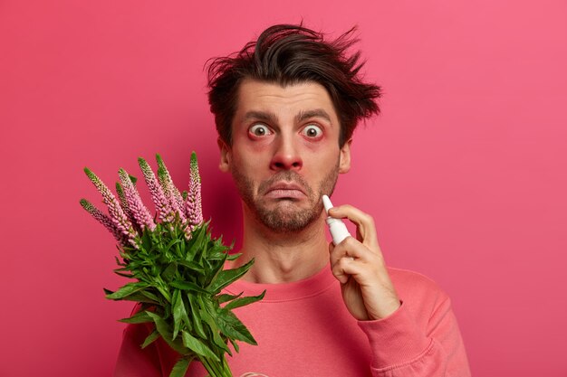 아픈 알레르기가있는 청년은 비강 방울로 코를 뚝뚝 떨어 뜨리고, 눈과 코가 빨갛게되며, 식물 알레르기, 비염 또는 건초열 증상, 응시, 분홍색 벽에 포즈를 취하고 꽃가루에 반응합니다.