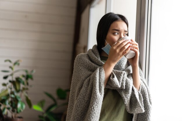 아픈 아시아 소녀가 집에서 격리된 채 뜨거운 차를 마시고 창밖을 바라보며 아프다...