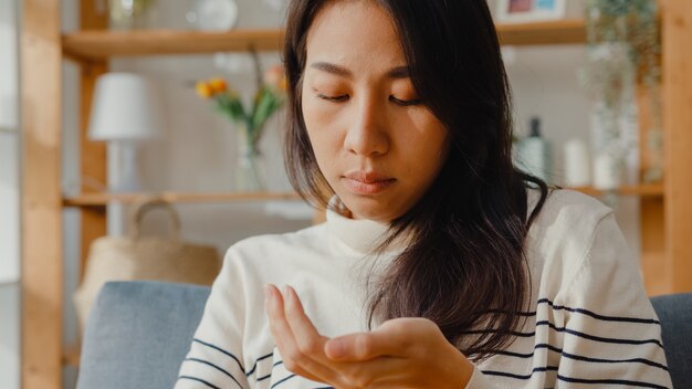 ピルを保持している病気のアジアの若い女性は、自宅のソファに座って探している薬を服用します