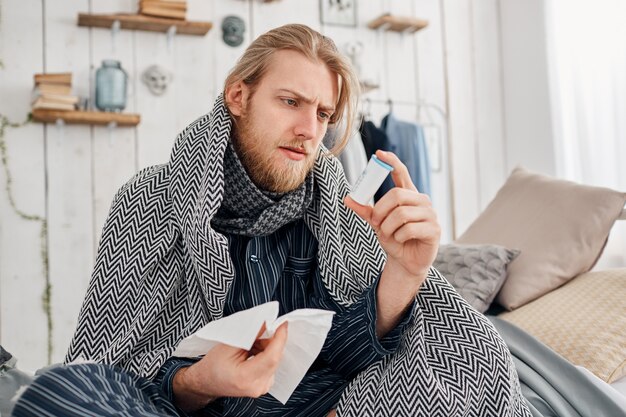 Sick bearded fair-haired man in sleepwear sits on bed surrounded by blanket and pillows, frowns while reading prescription on pills, holds handkerchief in hand. Health problems, bad cold and flu.